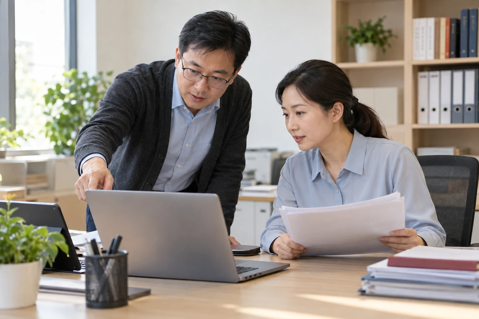 技术人员与教务人员在办公室交流人员同步资料,桌上设备清单无字,电脑背面可见,业务场景示意